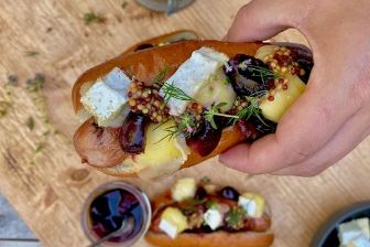 Gourmet frankfurter topped with creamy Mt Tam triple cream cheese, whole-grain mustard, pickled cherries, and fresh herbs, served in a toasted bun.