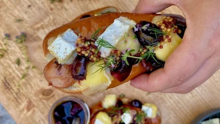 Gourmet frankfurter topped with creamy Mt Tam triple cream cheese, whole-grain mustard, pickled cherries, and fresh herbs, served in a toasted bun.